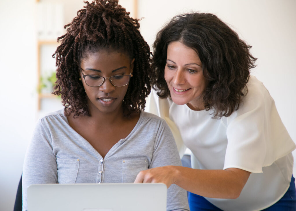 Two women looking at a laptop. One woman is pointing at the screen with her finger. The image represents an example of an in-person mentoring session that can be scheduled using our mentoring software.