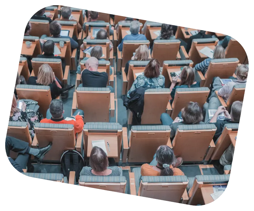 A view from above of a crowded lecture hall with rows of people seated in wooden chairs, some taking notes or using electronic devices. This setting reflects a professional or academic environment, possibly related to a seminar, workshop, or alumni event, where organisational tools like alumni management software might support event logistics and participant interaction.