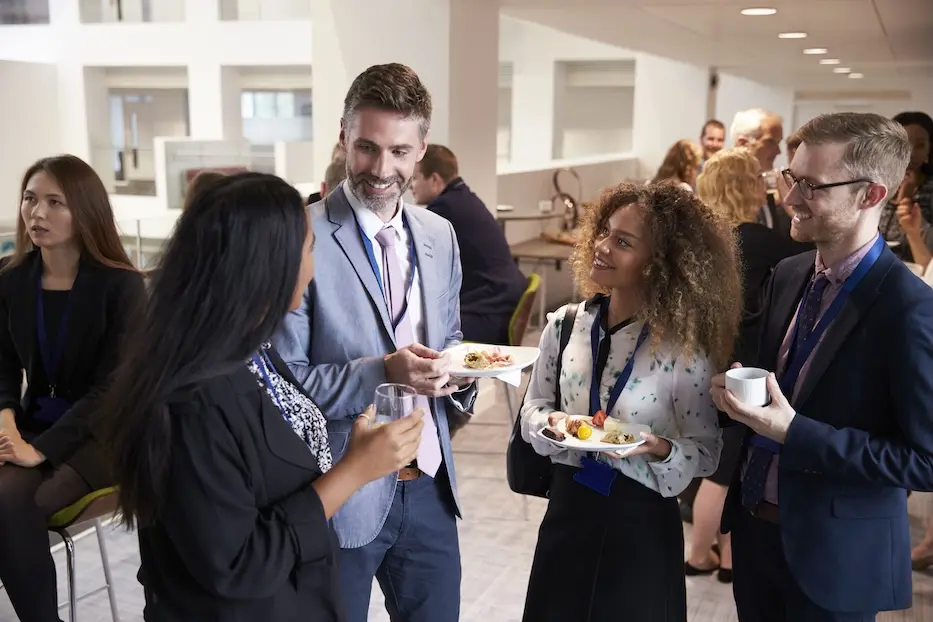 Group of professionally dressed people in conversation, holding plates of food. Represents event created using VeryConnect's alumni engagement platform.