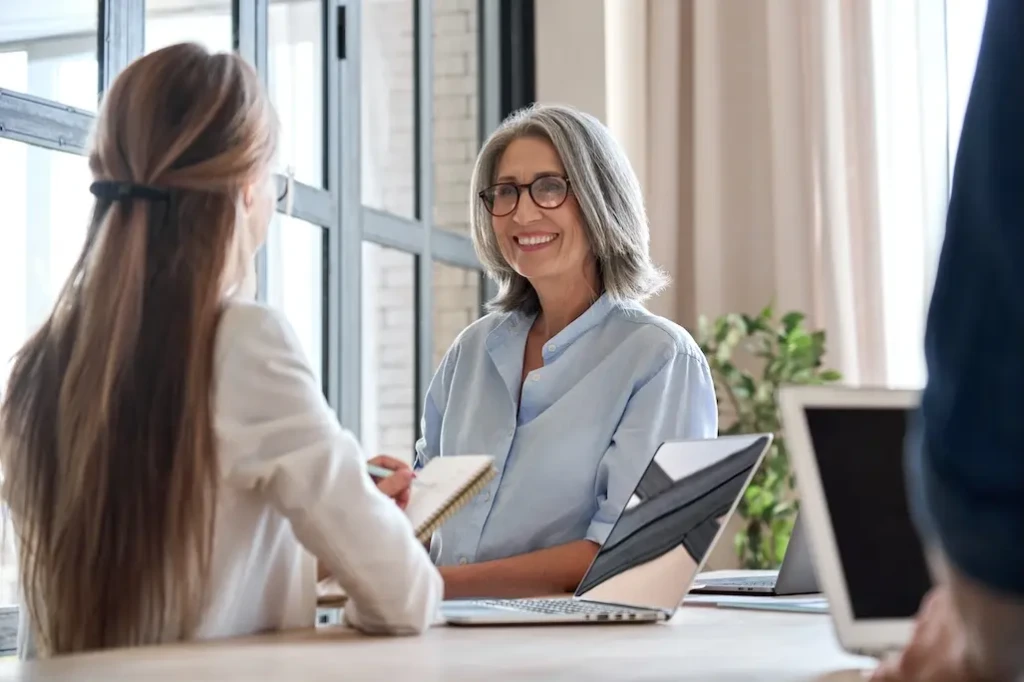 Two professionally dressed women. One sitting at laptop taking notes and the other smiling. Represents users of VeryConnect's alumni mentoring software.