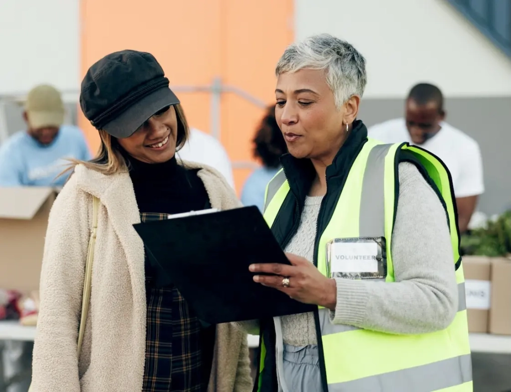 Two women, one with high-vis vest showing the other a clipboard. Represents user of VeryConnect's C charity management software