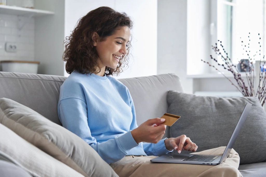 Woman sitting on couch with payment card in hand, looking at laptop. Represents user of donation management software.