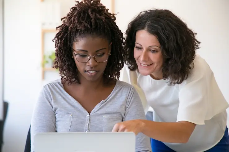 Two women looking at a laptop. One woman is pointing at the screen with her finger. The image represents an example of an in-person mentoring session that can be scheduled using mentoring software.